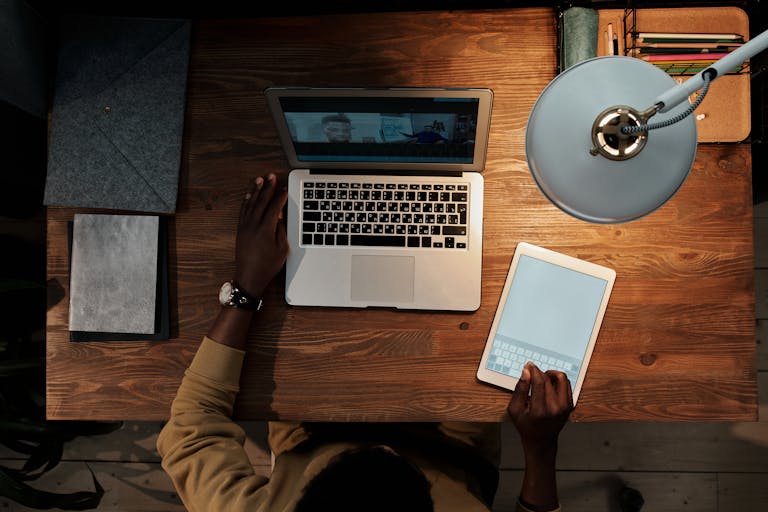 Person using a tablet while working on a laptop during a video call, viewed from above.